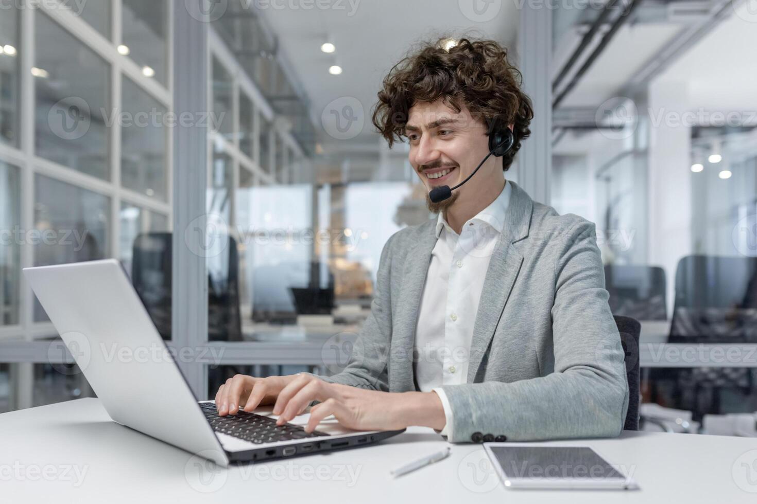 sonriente joven empresario con auriculares trabajando en ordenador portátil en moderno oficina, exhibiendo multitarea y cliente Servicio habilidades. positivo trabajo ambiente reflejado en su alegre expresión. foto