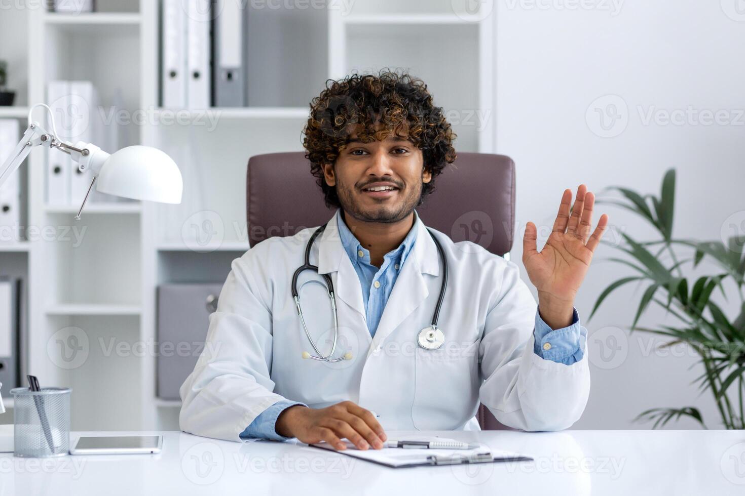 Webcam view, online call consultation, young Indian doctor in white medical coat looking at camera, waving hand greeting to patient, doctor working remotely inside clinic office with laptop. photo