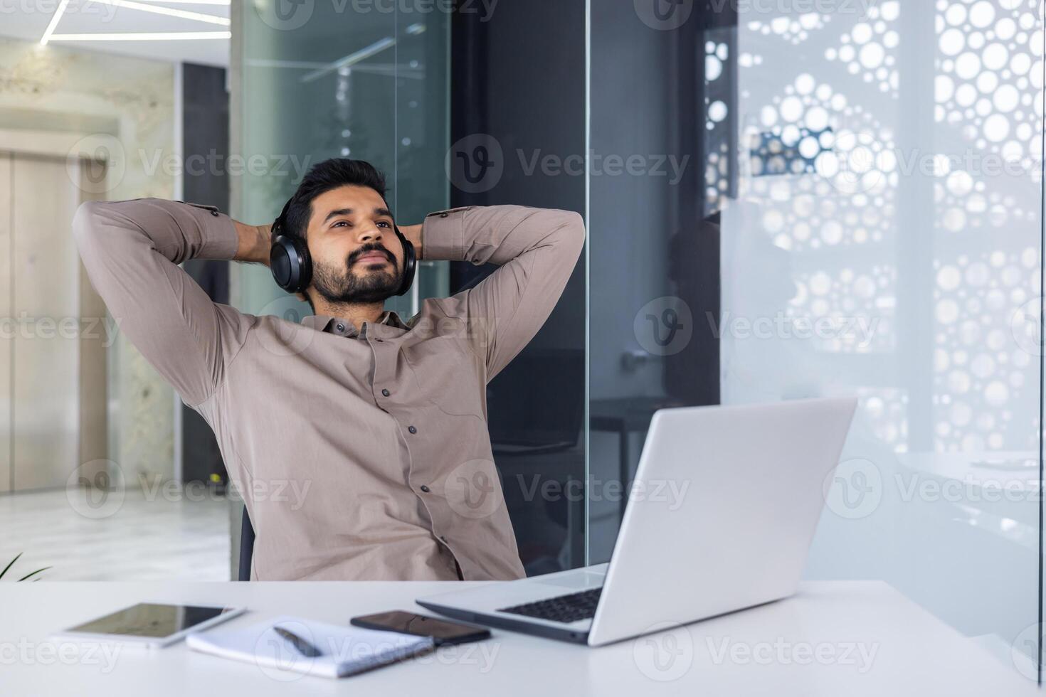 Indian young man relaxing at the workplace, satisfied with the finished project and the result. He sits relaxed at the table, wearing headphones, with his hands behind his head. photo