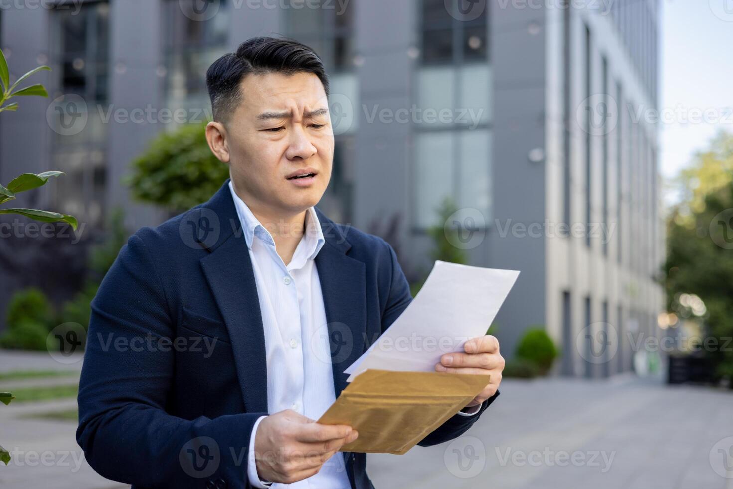 Upset and disappointed Asian businessman sitting outside the office and reading a received letter with bad news. photo