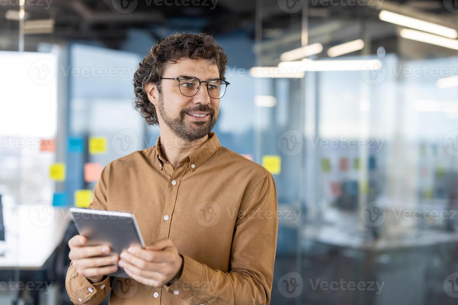 Confident male with curly hair looking aside with curious face while keeping electronic device in hands. Professional manager watching as colleagues passing by in hall of modern office. photo