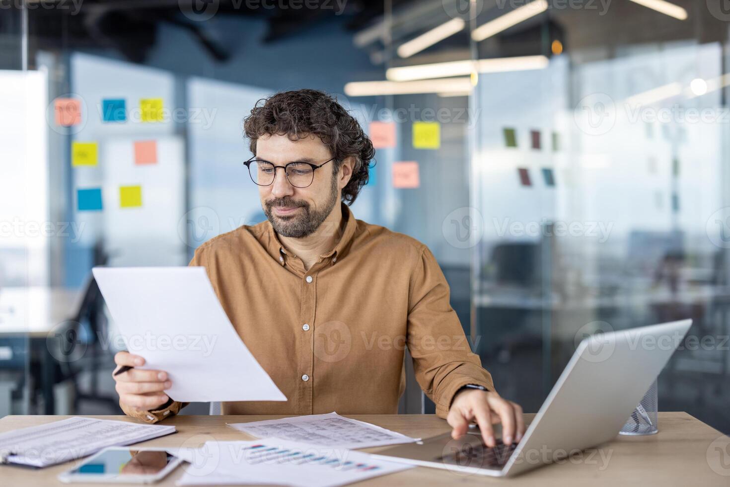 A focused male business professional examines papers while seated at a desk with a laptop in a contemporary office setting, portraying productivity and dedication. photo