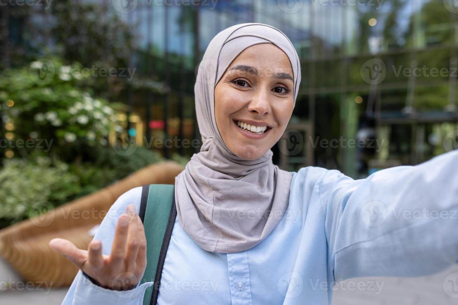 A cheerful student in a hijab takes a selfie, possibly on a call, amidst a vibrant cityscape. photo