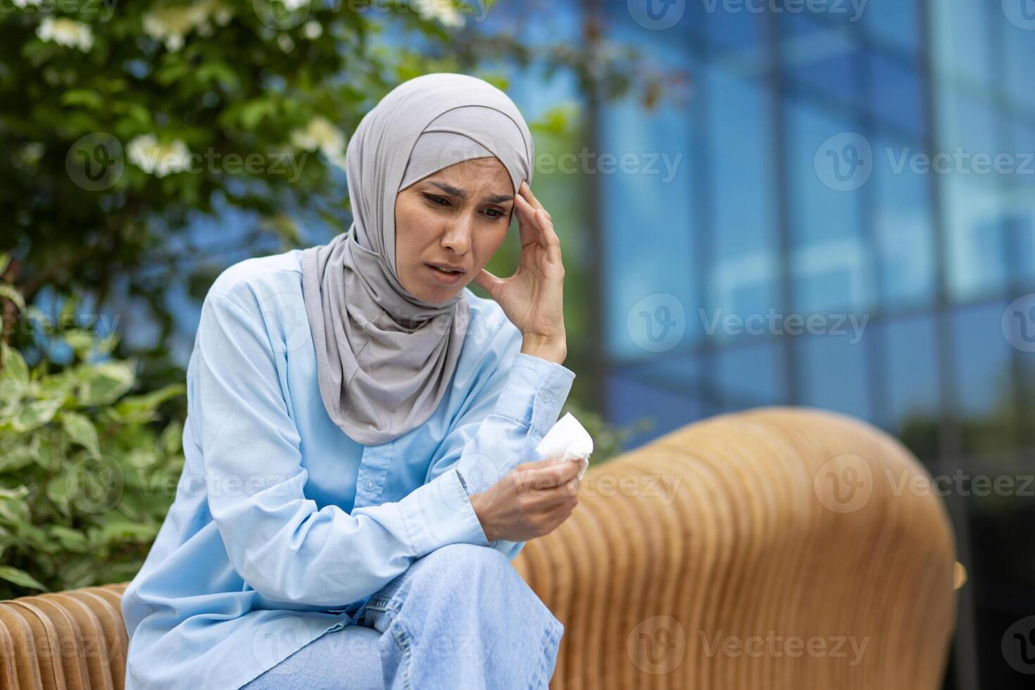 A middle-aged woman wearing a blue hijab looks distressed and unwell, sitting outdoors with tissues in her hand, expressing discomfort or illness in an urban setting. photo