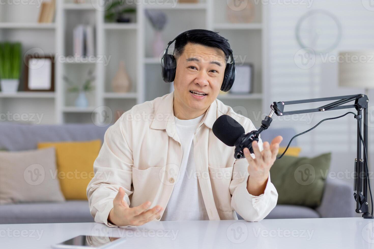 Friendly Asian man with headphones podcasting, talking and gesturing towards the camera with a broadcast microphone and a laptop in a home studio setting. photo