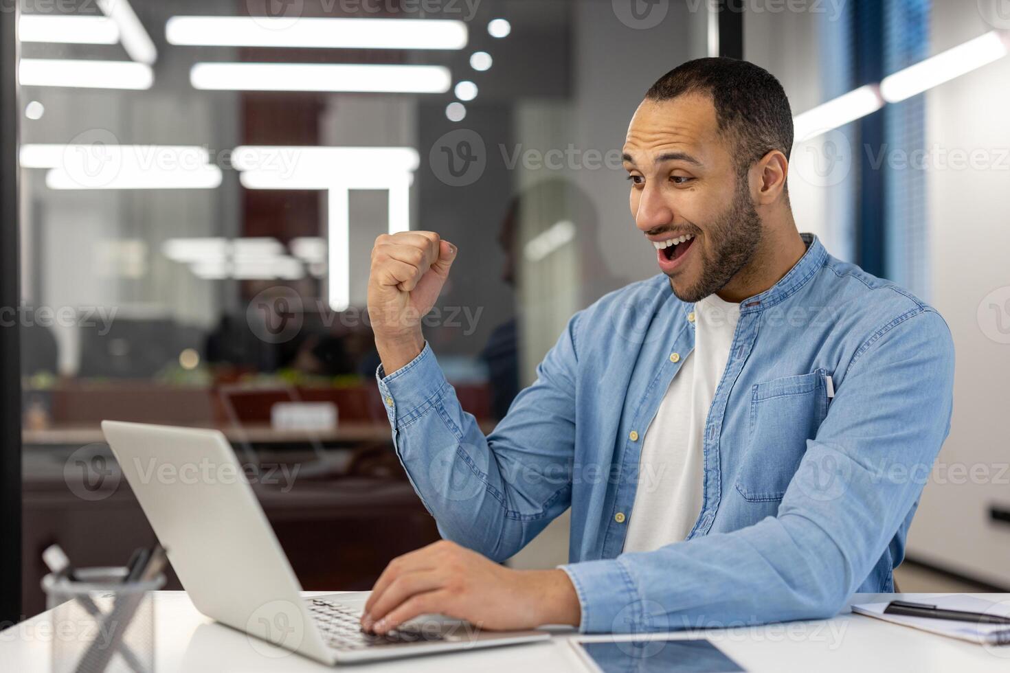Happy young hispanic man sitting in modern office at work desk and looking surprised at laptop screen, happy with result and news, showing victory gesture yes with hand. photo