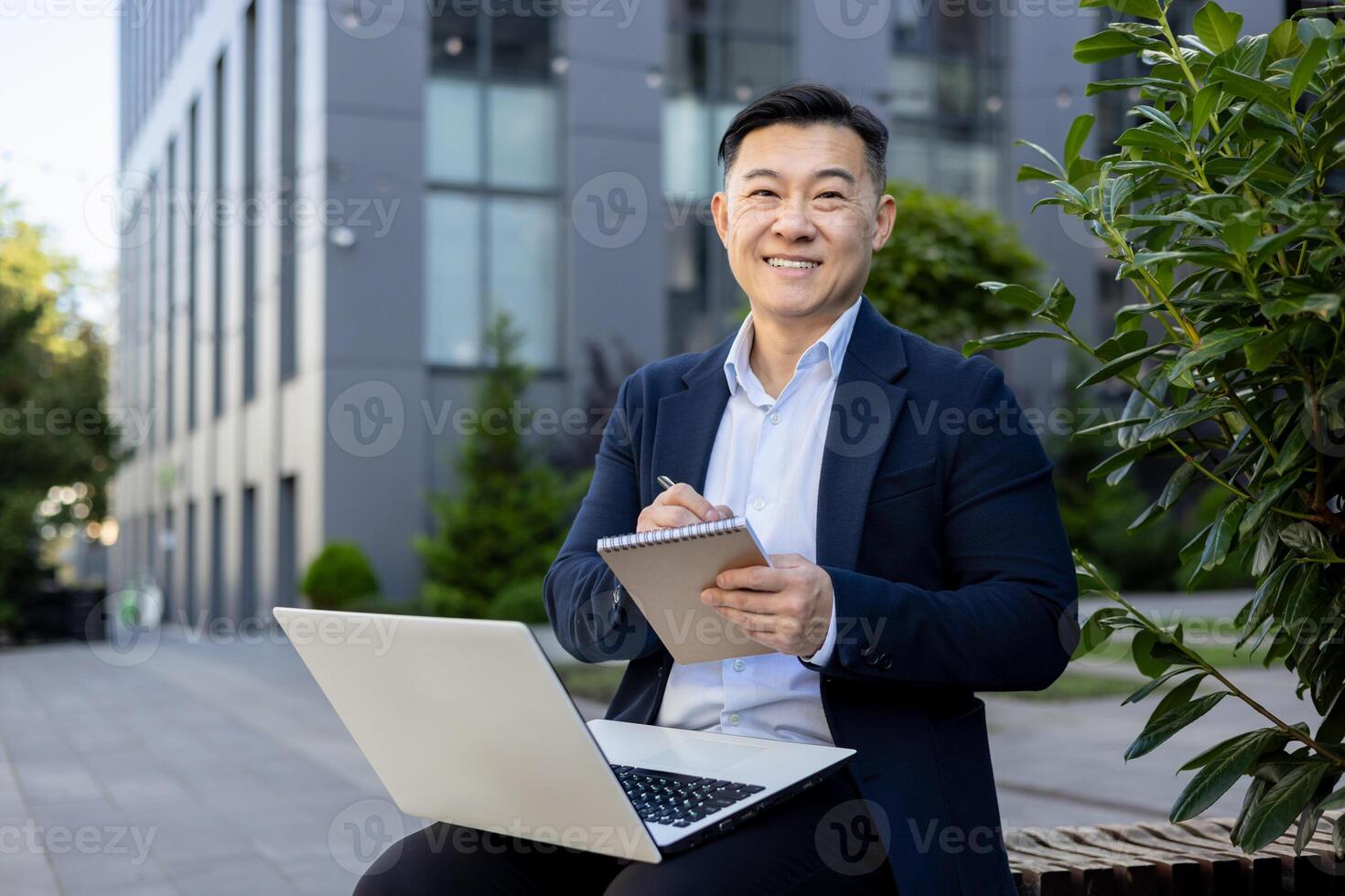 Portrait of a successful and smiling young Asian man in a business suit sitting outside a business center, holding a laptop on his lap and a pen with a notepad in his hands, looking at the camera. photo