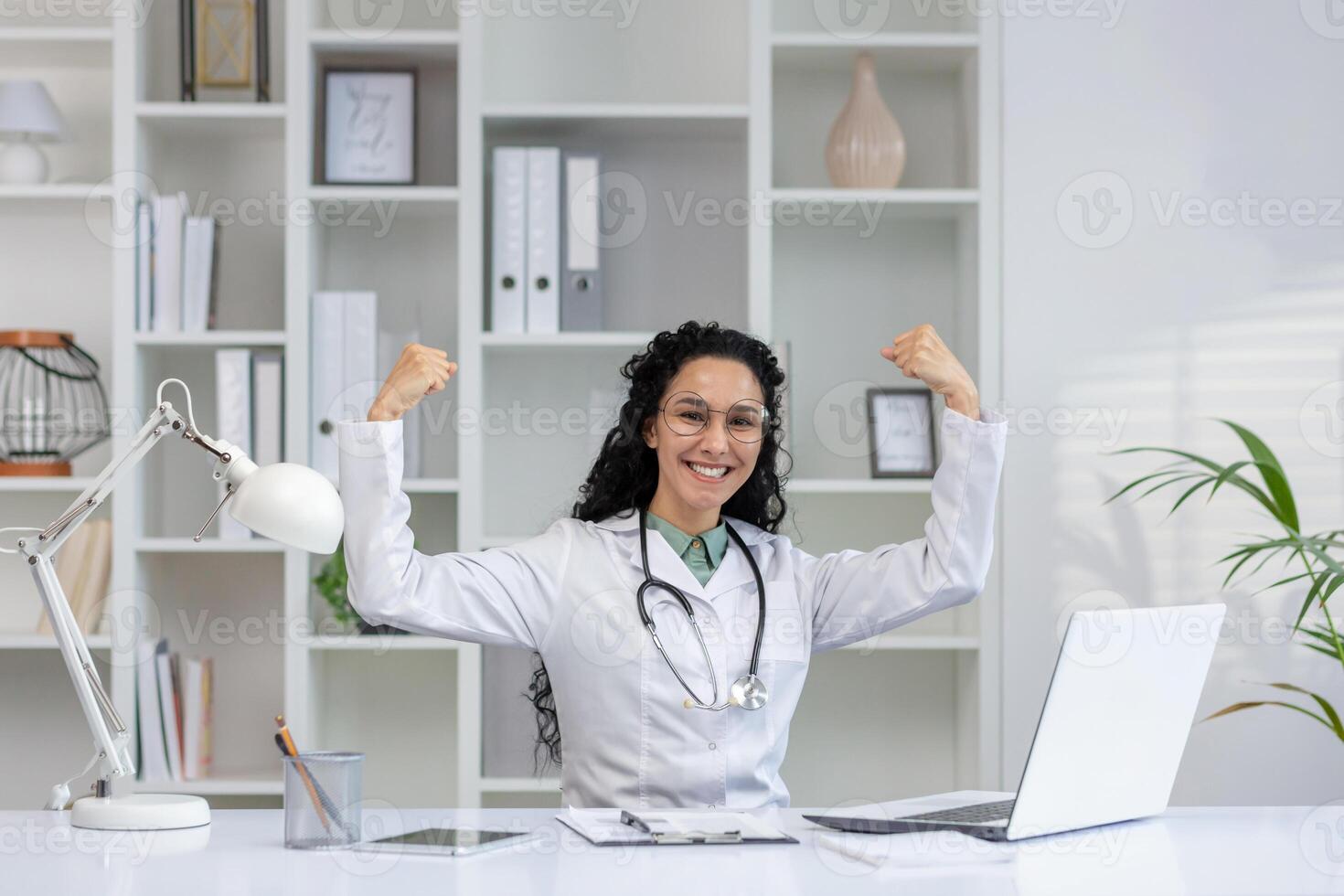 A joyful latino woman doctor with curly hair raises her arms in a triumphant pose, celebrating a professional success in her medical office. photo