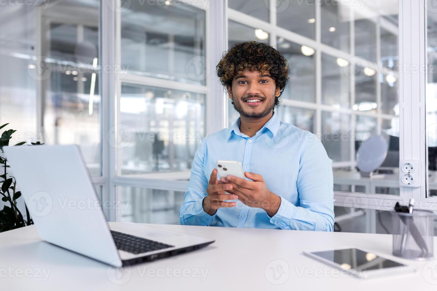 A young Latin American businessman sits in the office at a desk and uses a mobile phone, writes a message, searches for information on the Internet, checks social networks. Smiling at the camera. photo