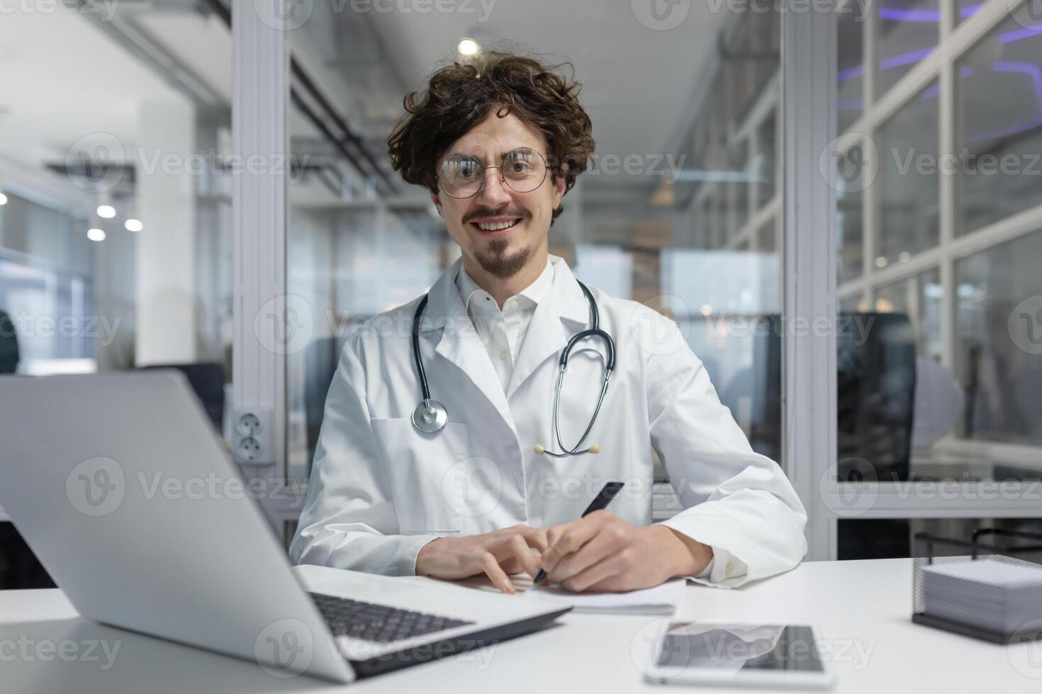 A doctor wearing a white lab coat and stethoscope is seated at a desk, using a laptop.Man smile happy writing report looking camera photo