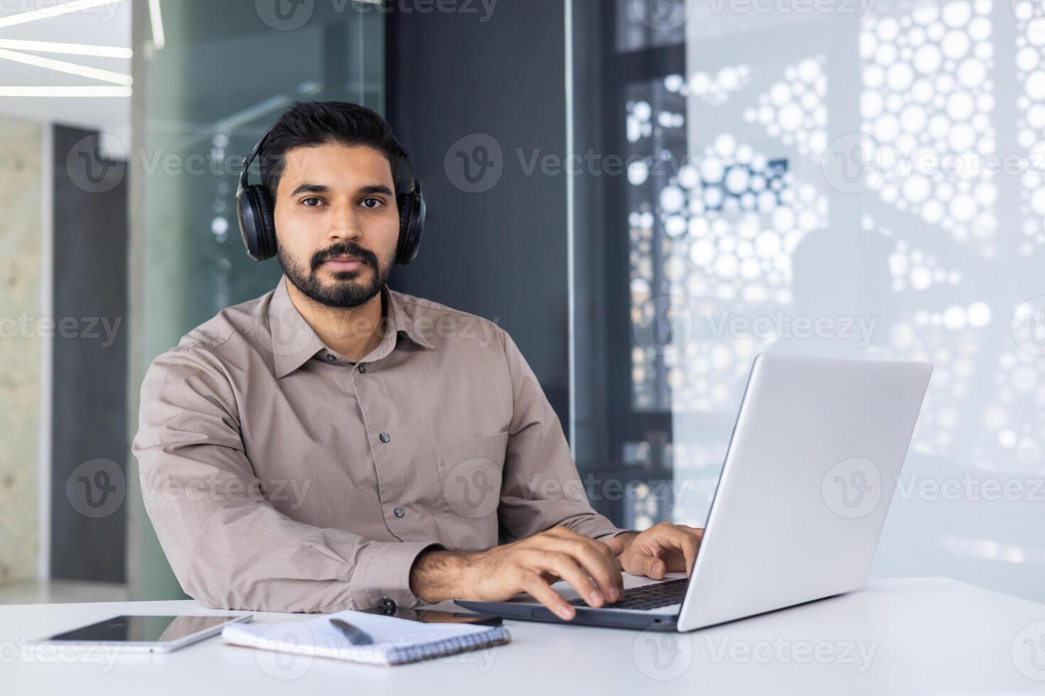 retrato de un grave joven indio hombre empresario, persona de libre dedicación trabajando en el oficina con un ordenador portátil. sentado a el mesa en auriculares y mirando a el cámara. foto