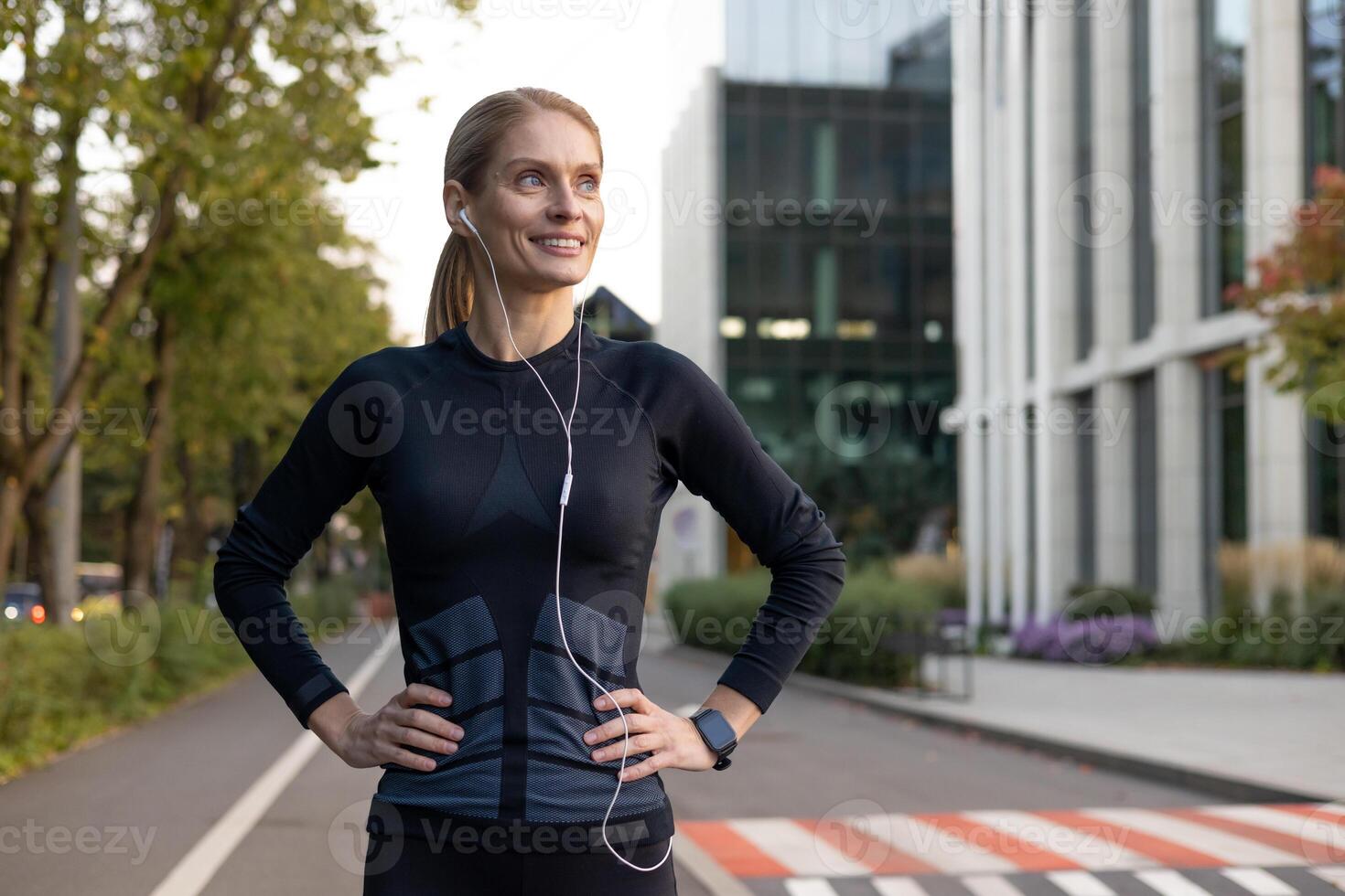 A fit and happy woman takes a break from running, her hands on her hips as she pauses to enjoy the city scenery. Earphones in, she's dressed in athletic gear, ready to continue her fitness routine. photo