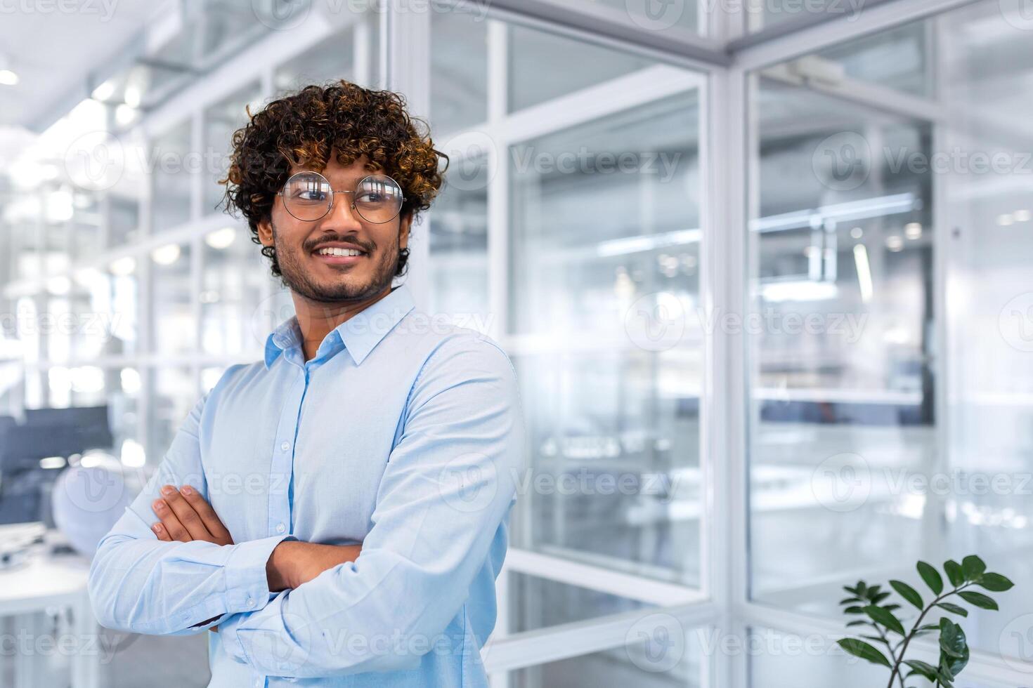 Portrait of young successful businessman inside modern office, hispanic man satisfied smiling and thinking, crossed arms and blue shirt and glasses looking window, good financier achievement photo