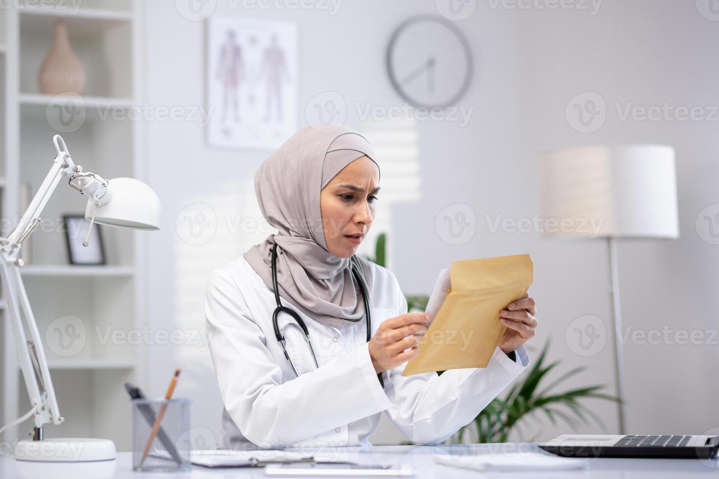 Disturbed female nurse in hijab opening paper envelope while sitting at workplace. Disappointed hospital worker reading unpleasant message from laboratory with unsuccessful treatment results. photo