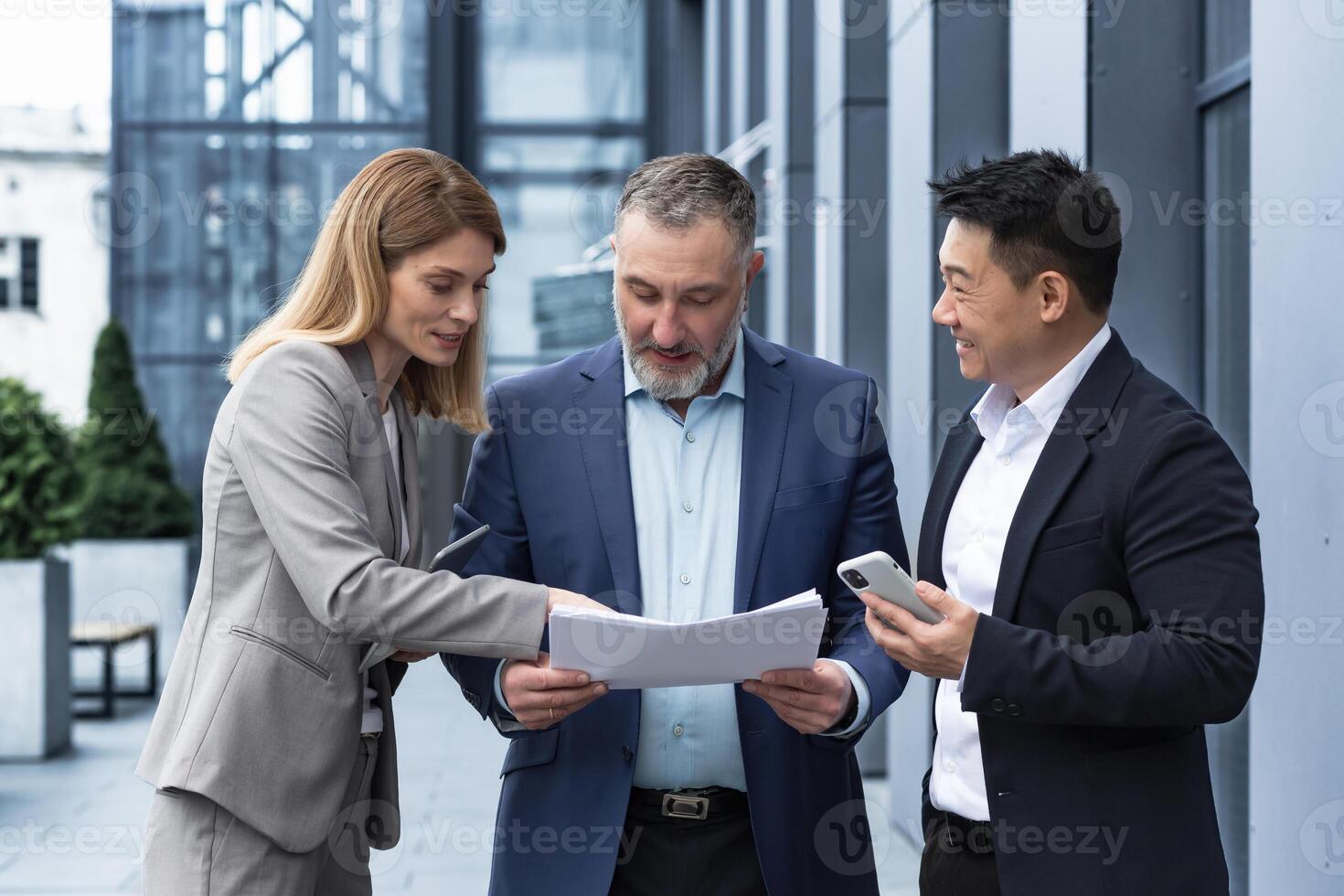 Three businessmen outside office building with documents discussing plans, businessmen and businesswoman having fun chatting and talking, diverse business group in business suits photo