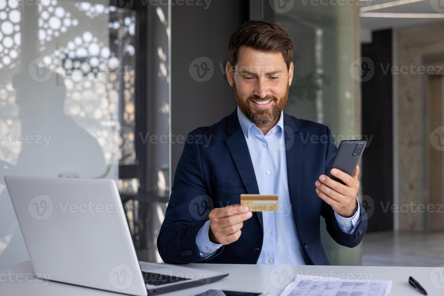 Smiling young businessman sitting in office at desk, holding phone and credit card, conducting online money transactions, transferring funds, checking account. photo