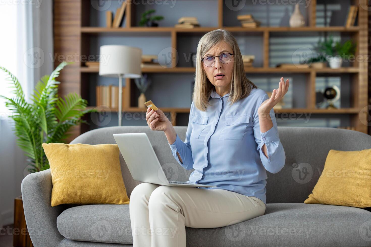 Mature woman at home looking at credit card with confusion, sitting on couch with her laptop, in a modern living room setting. photo