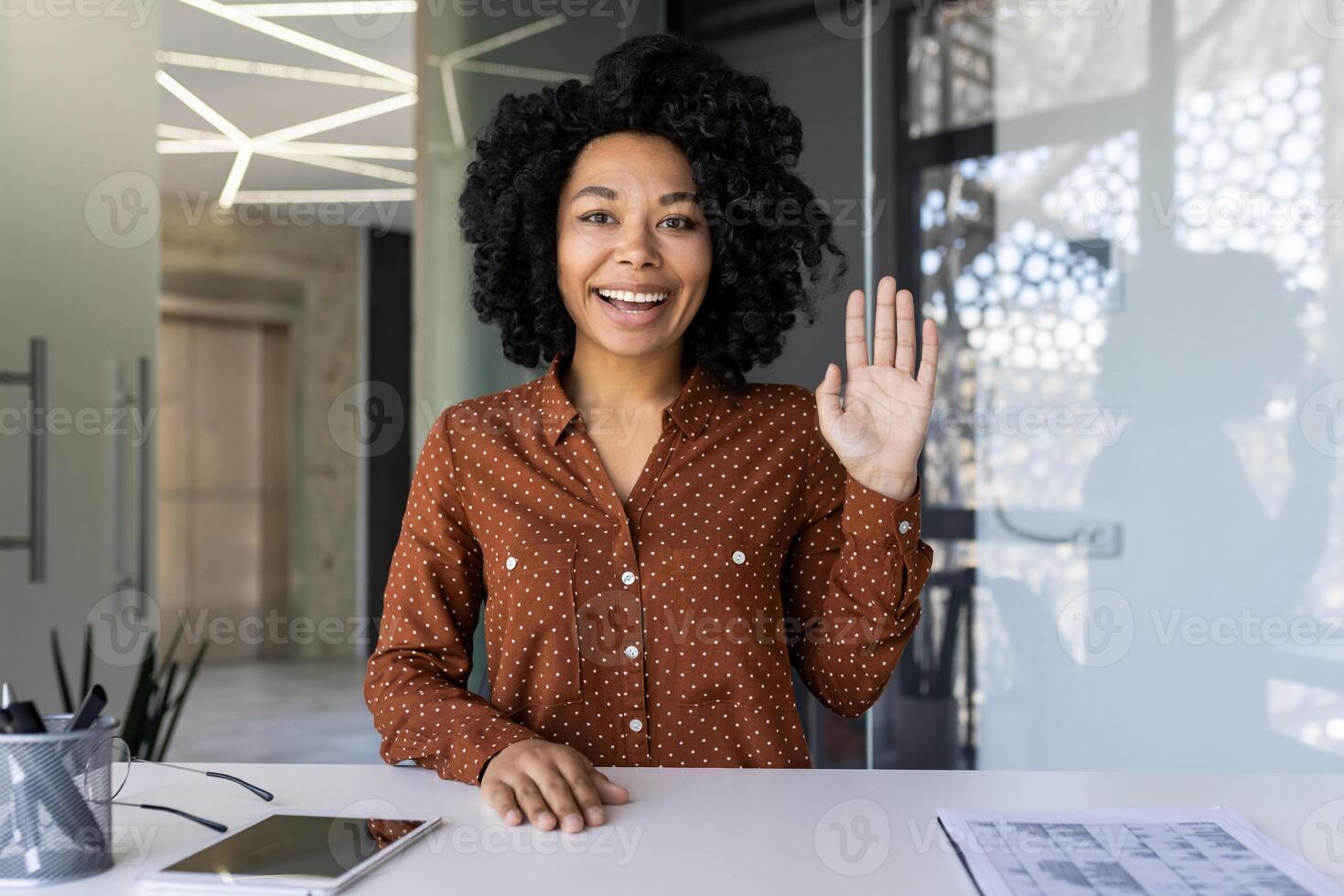 Cheerful young woman engaging in a call, waving to the camera with a big smile, well-lit modern office background. photo