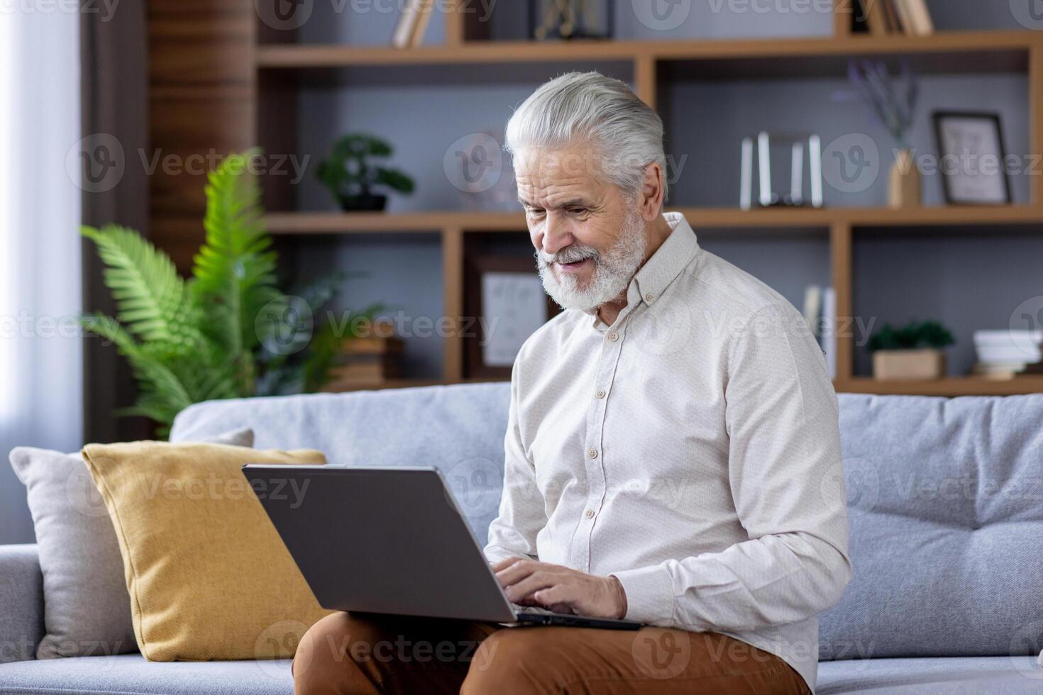 un alegre mayor hombre con gris pelo se sienta cómodamente en un sofá, profundamente comprometido en utilizando su ordenador portátil dentro un moderno vivo habitación decorado con interior plantas. foto