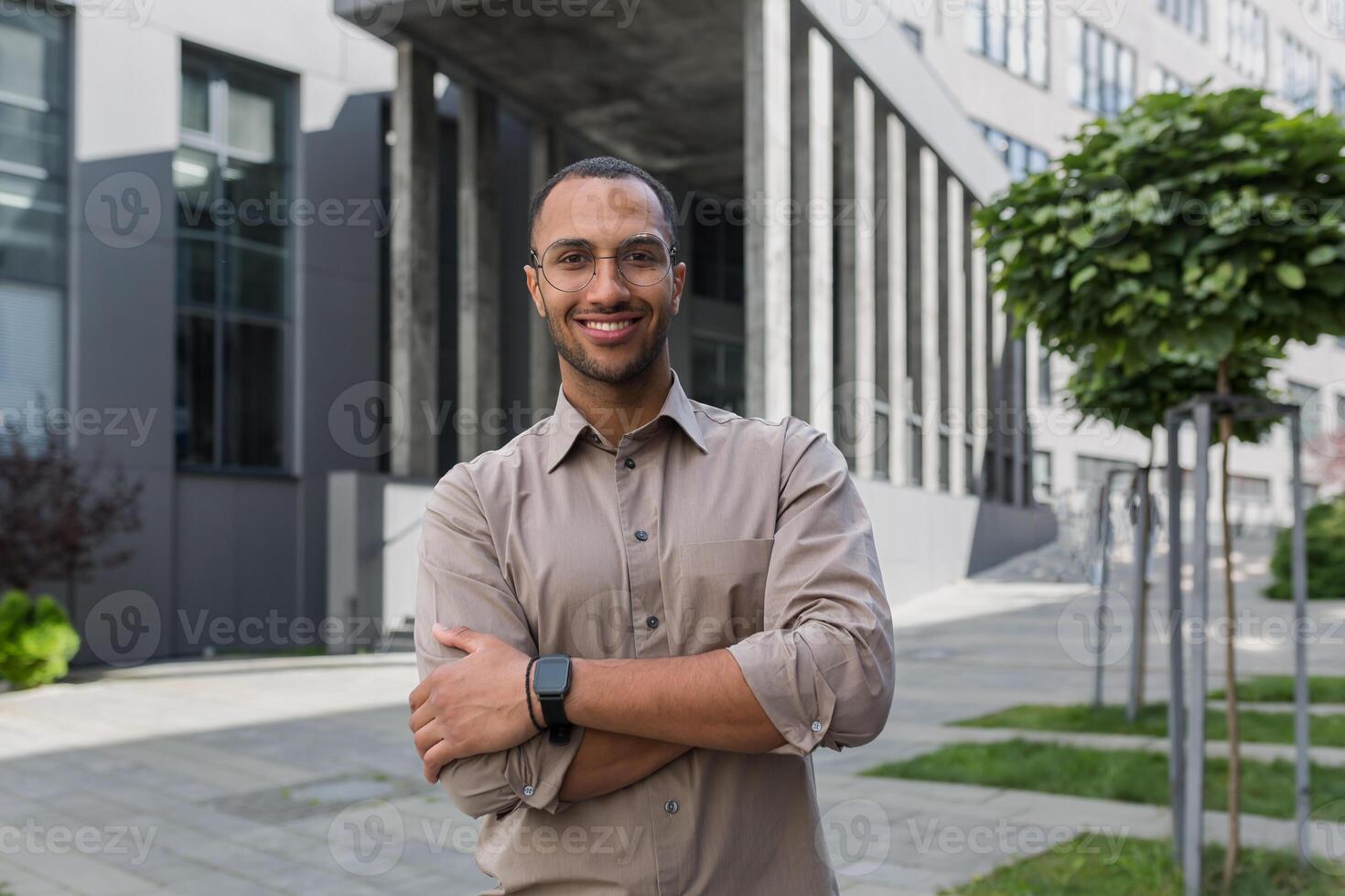 joven exitoso empresario sonriente y mirando a cámara con cruzado brazos, hombre fuera de oficina edificio vistiendo camisa y anteojos, puesta en marcha inversor foto