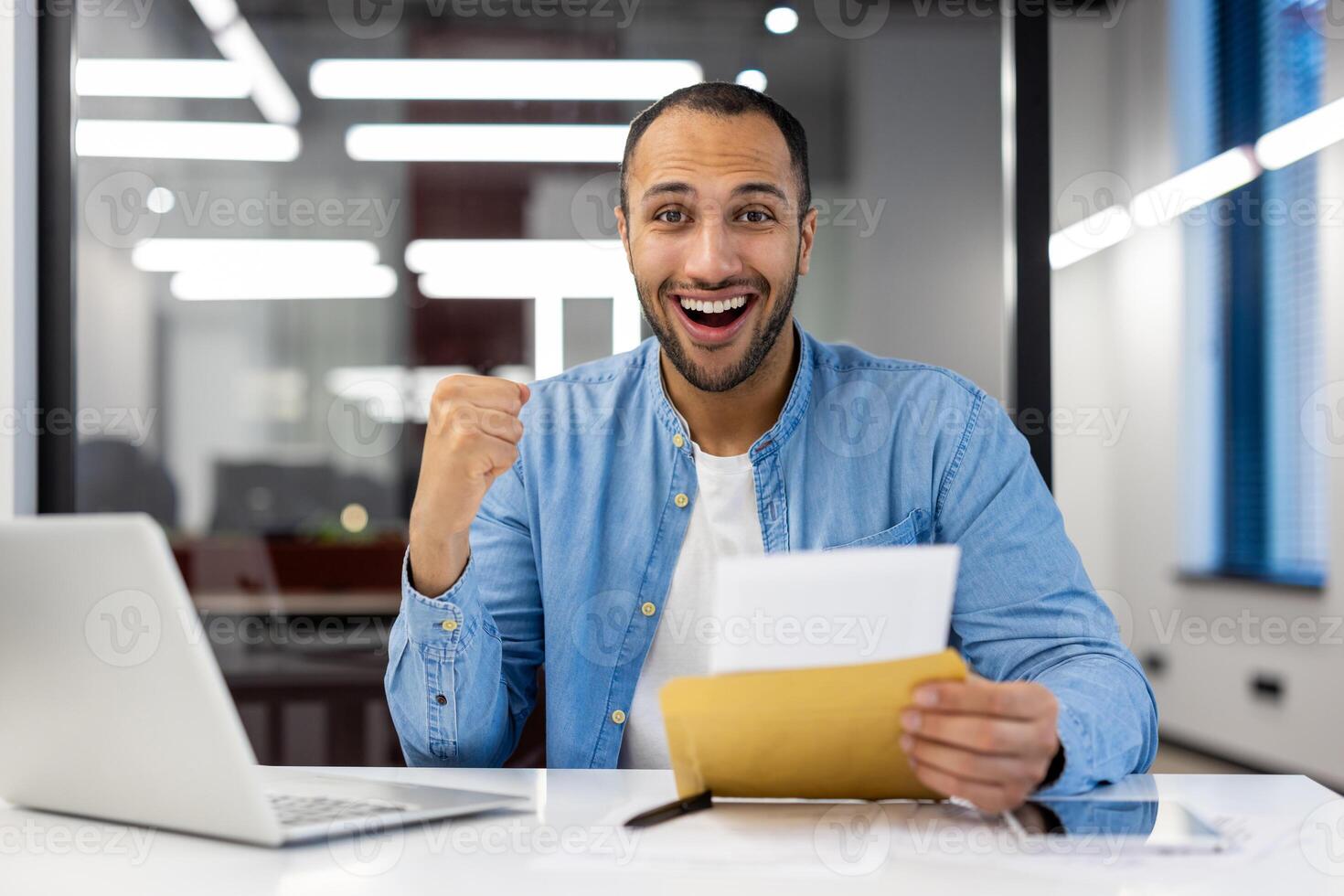 Portrait of a sad young Muslim man sitting in the office at the desk, holding an open envelope with a letter in his hands and looking happy at the camera, showing a victory gesture with his hand. photo