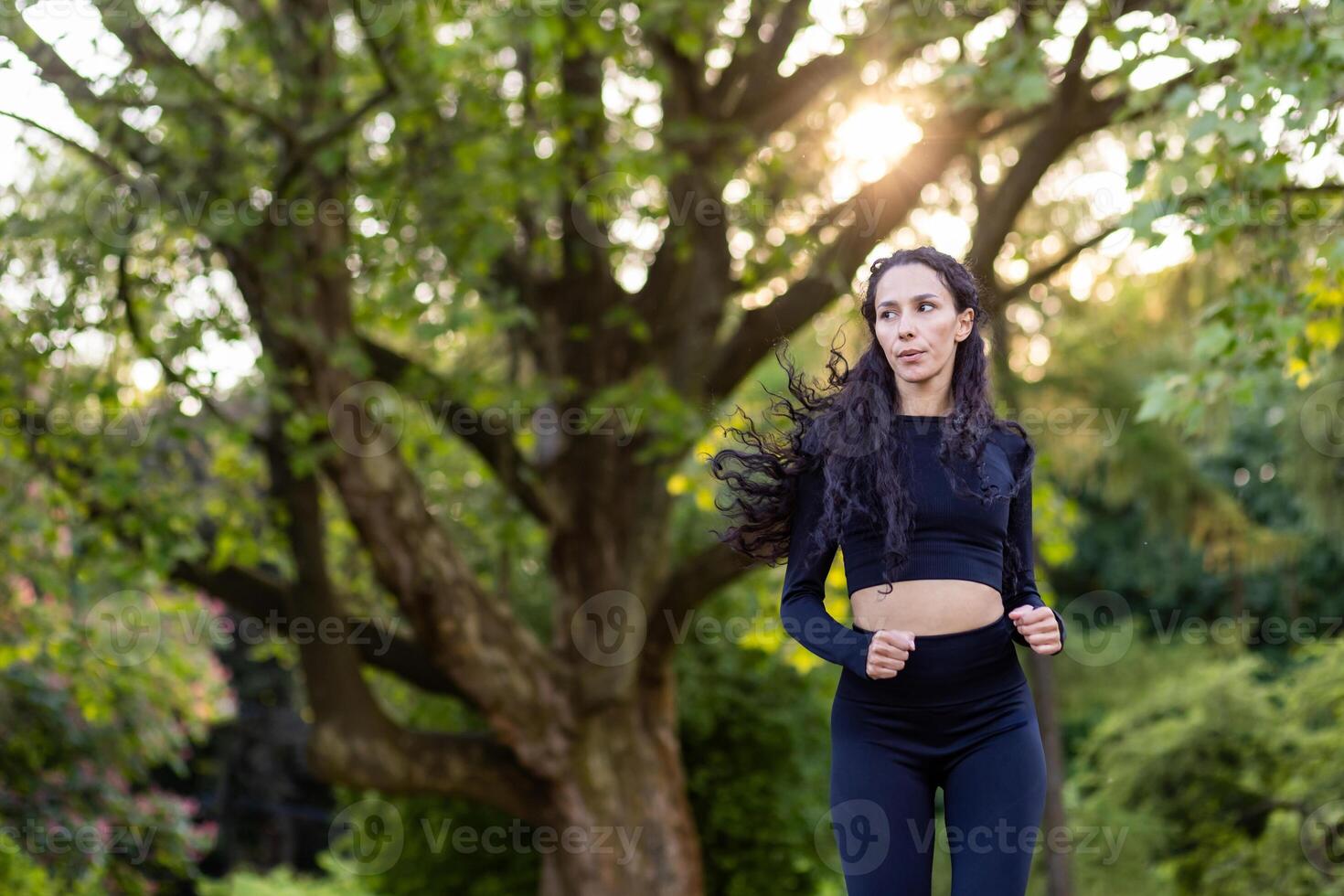 Beautiful and slim Hispanic woman in tracksuit running in park, woman with curly hair among trees. photo
