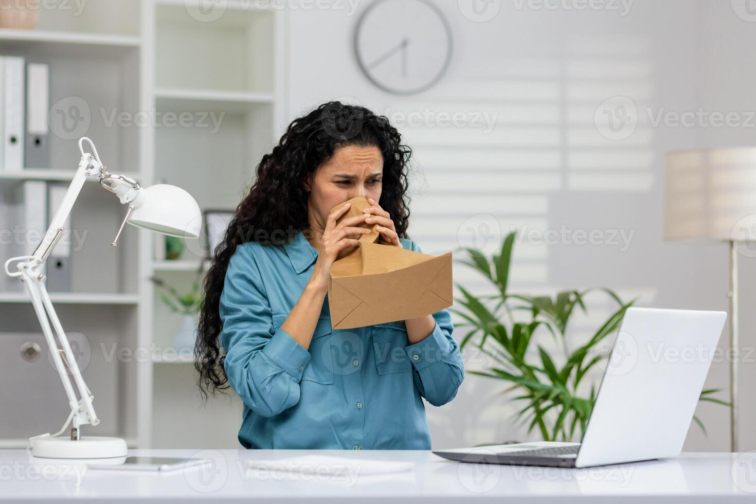 A distressed businesswoman in a blue shirt is breathing into a paper bag at her workplace, showing signs of a panic attack with a laptop in front of her. photo