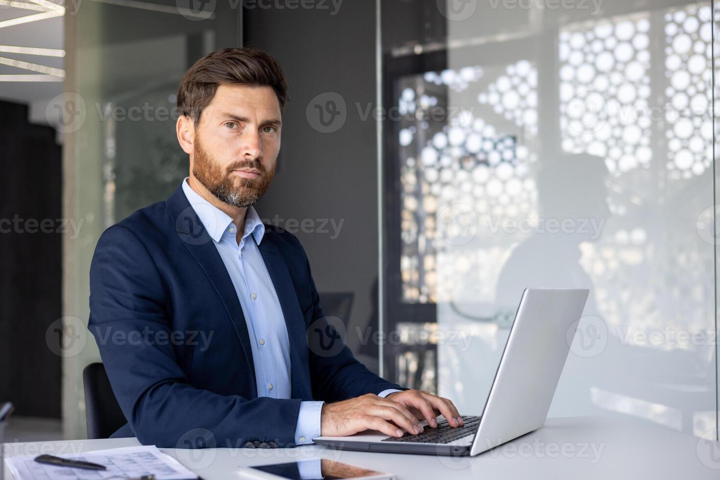 Portrait of a serious and self-confident young businessman sitting in the office at the table, typing on a laptop, working with clients and documents, looking at the camera. photo