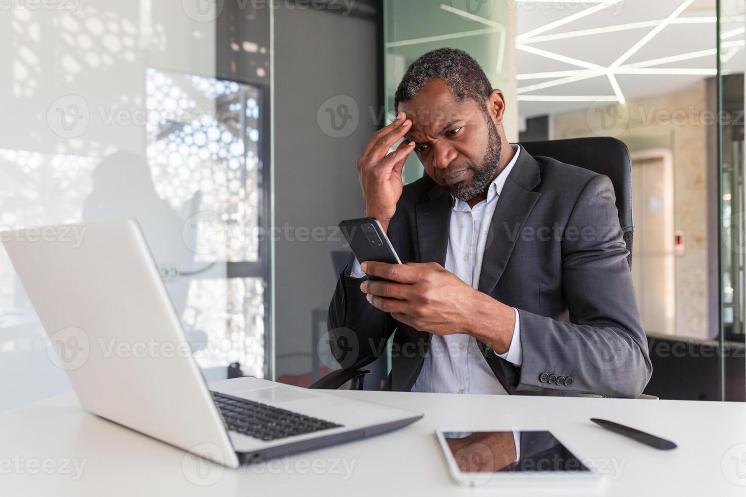 Upset and disappointed man inside office holding phone, businessman reading bad news at workplace inside office, african american senior boss unhappy with achievement results notification. photo