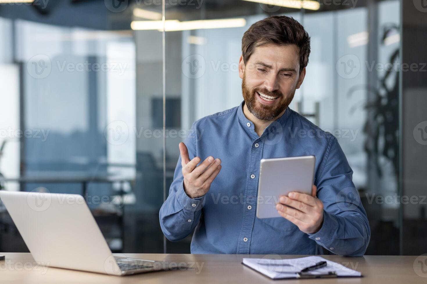 A young man sits in the office at a desk with a laptop and smilingly talks on a call on a tablet. photo