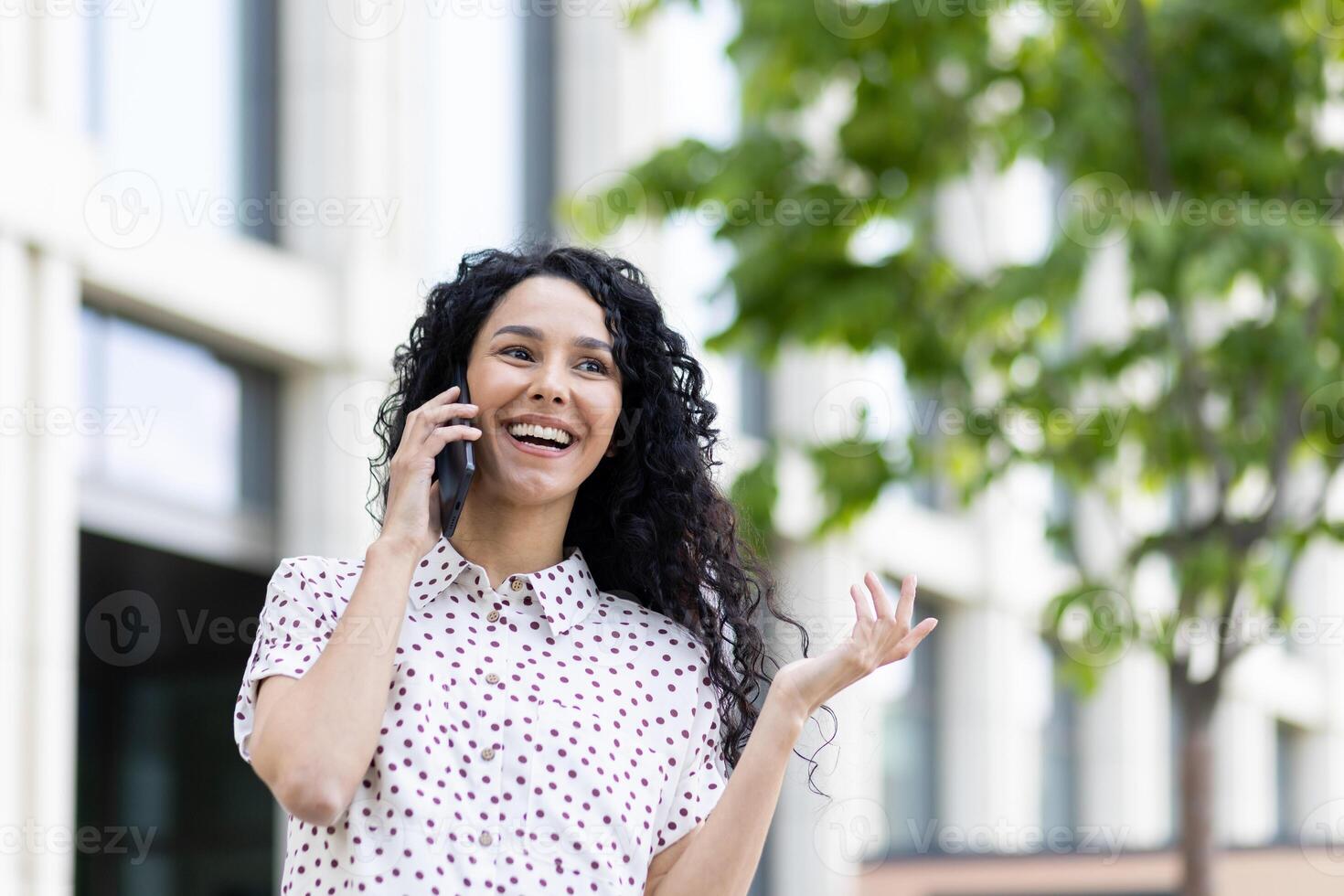 Young beautiful woman talking on the phone close-up while walking in the city, business woman smiling contentedly, successful Latin American woman with curly hair on a sunny day. photo
