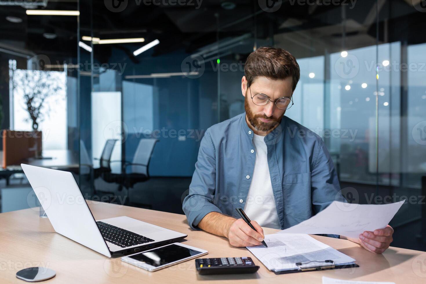 Serious and focused financier accountant on paper work inside office, mature man using calculator and laptop for calculating reports and summarizing accounts, businessman at work in casual clothes. photo