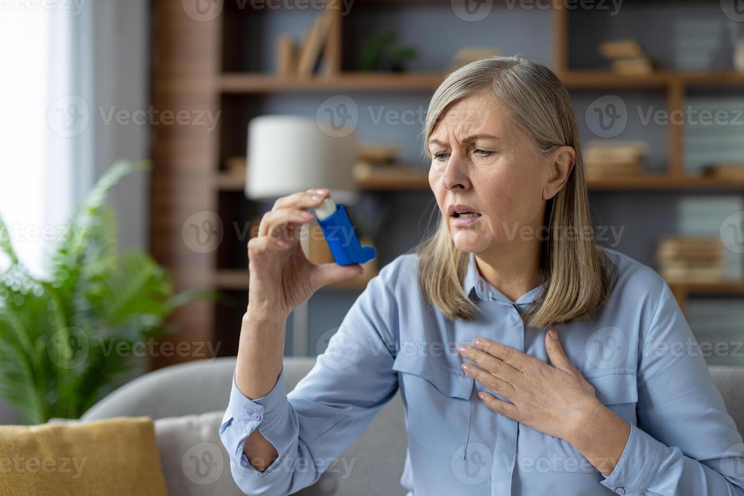 An elderly woman experiences an asthma attack and uses a blue inhaler. She looks concerned while sitting in a modern living room. photo