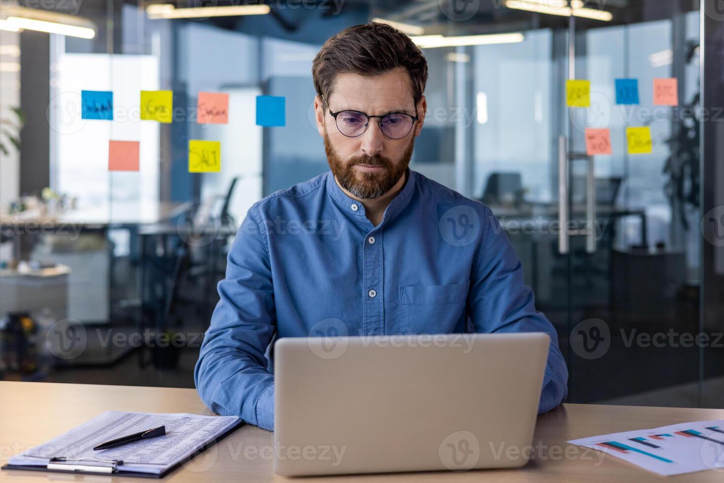 retrato de un grave y enfocado joven especialista, desarrollador y programador sentado en el oficina a el mesa y trabajando en un ordenador portátil. foto