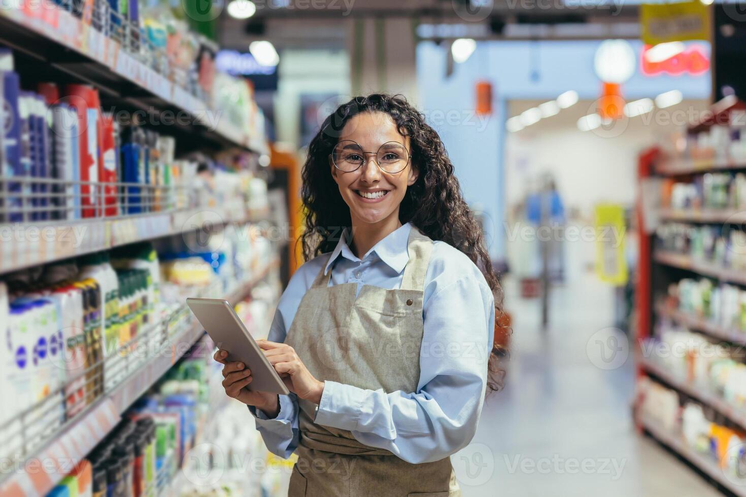 Portrait of happy and successful saleswoman, hispanic woman with curly hair smiling and looking at camera, using tablet computer to review product photo