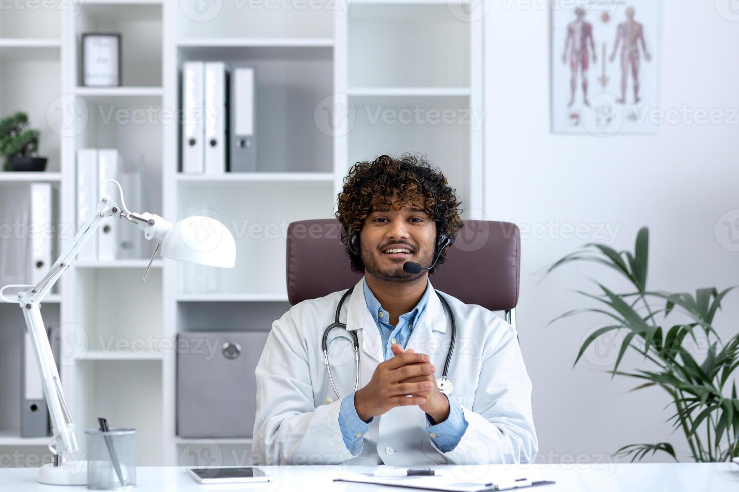 Webcam view, young doctor with headset phone using laptop for call, doctor cheerfully and friendly consulting patients, smiling and looking at camera, working inside clinic medical room. photo