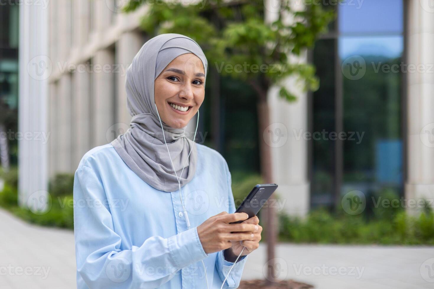 Smiling young Muslim female professional in hijab engaging with smartphone in front of an office building in the city. photo