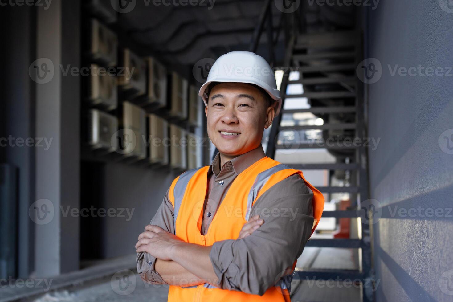 retrato de un sonriente asiático masculino ingeniero, construcción obrero, técnico en pie fuera de un edificio en un difícil sombrero y chaleco, cruce su brazos terminado su cofre y mirando con confianza a el cámara. foto