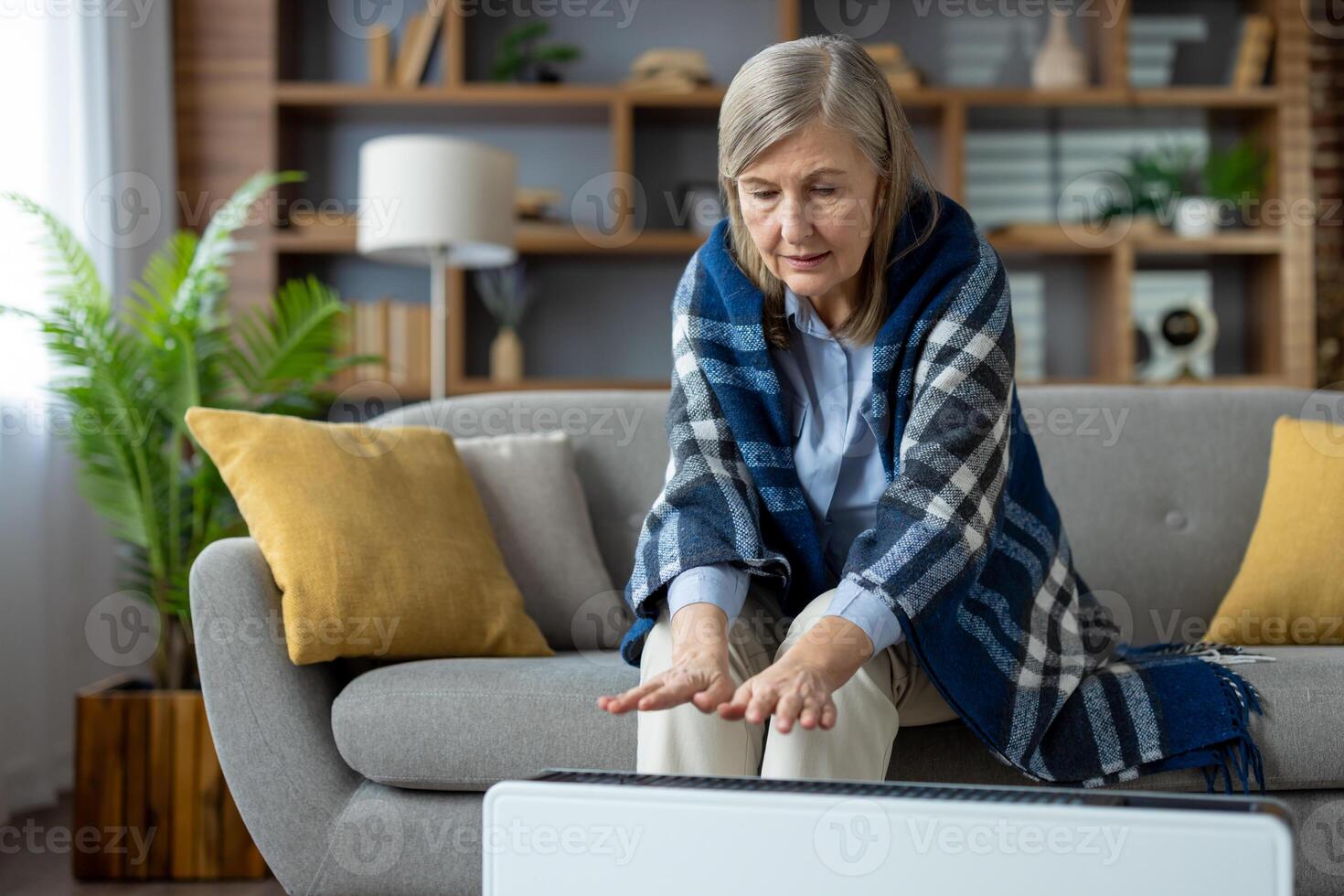 Blonde mature woman under wool coverlet sitting on couch and stretching hands to electric radiator. Caucasian female trying getting warm in spacious apartment during central heating off season. photo