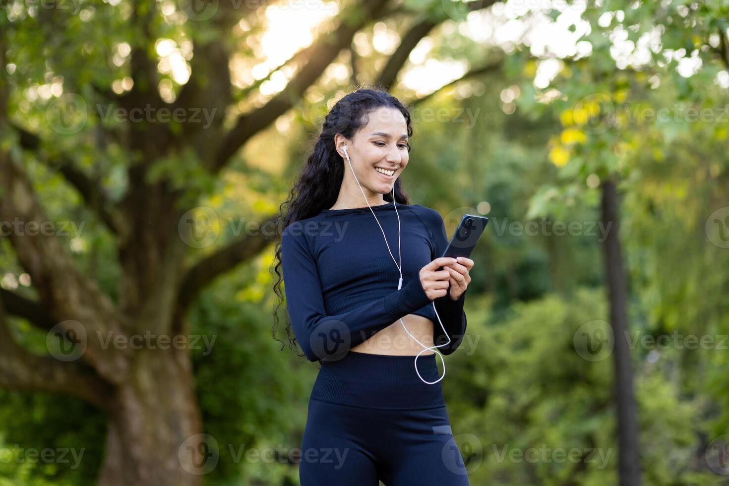 Fitness enthusiast takes a break in a lush park to choose music or track progress on her phone, capturing a moment of technology in fitness. photo