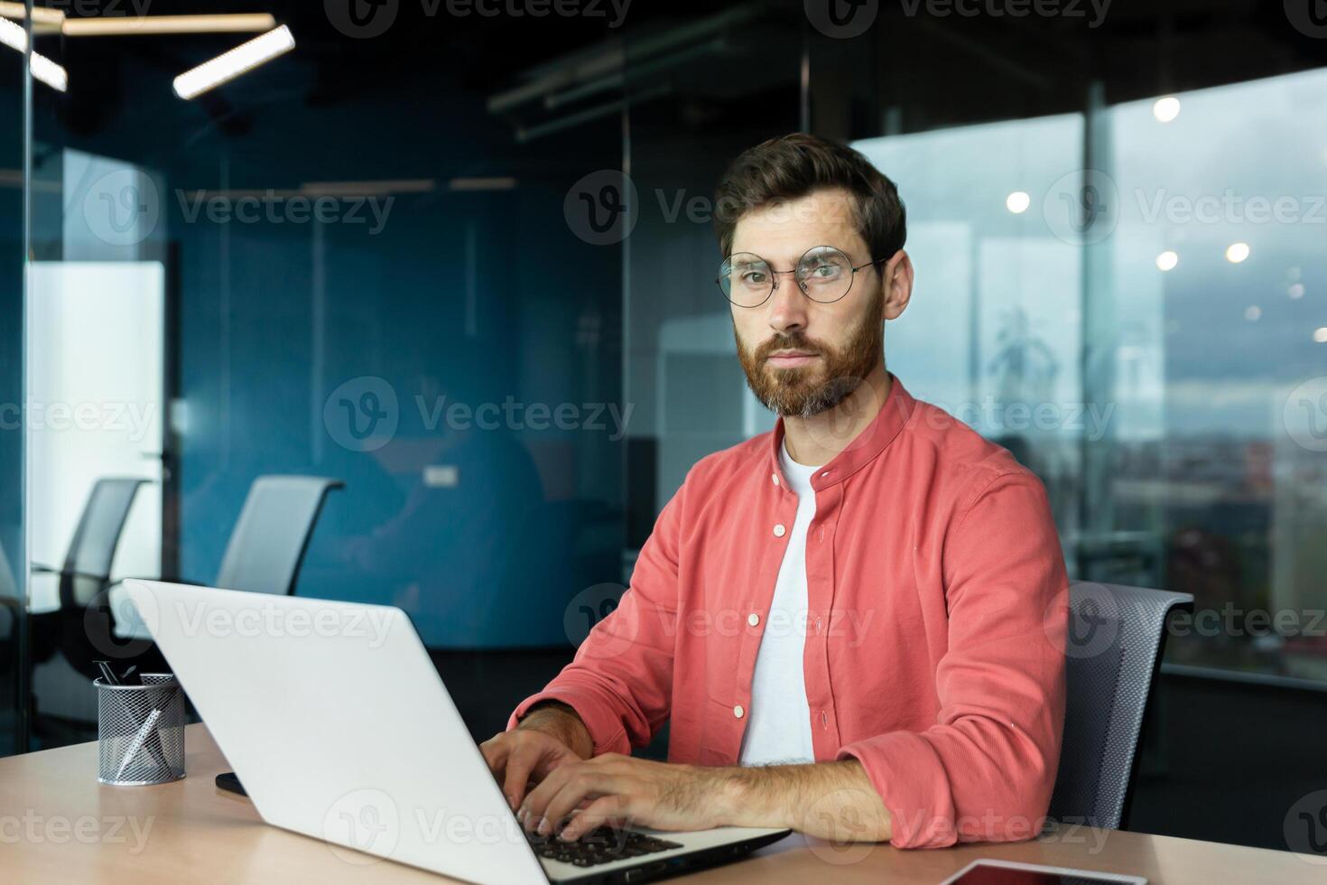 Serious and focused mature businessman in red shirt wearing glasses looking at camera, young man at work typing on laptop keyboard, programmer developer portrait at work. photo