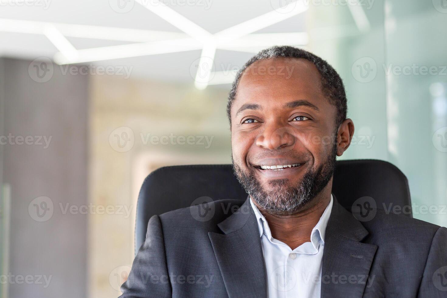 Close up of successful dreamy businessman smiling and looking at camera, african american boss in business suit smiling and looking at window inside office. photo