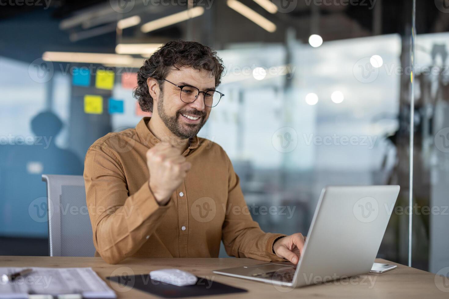 alegre empresario con lentes celebrando un éxito mientras trabajando en su ordenador portátil en un moderno oficina ambiente. foto