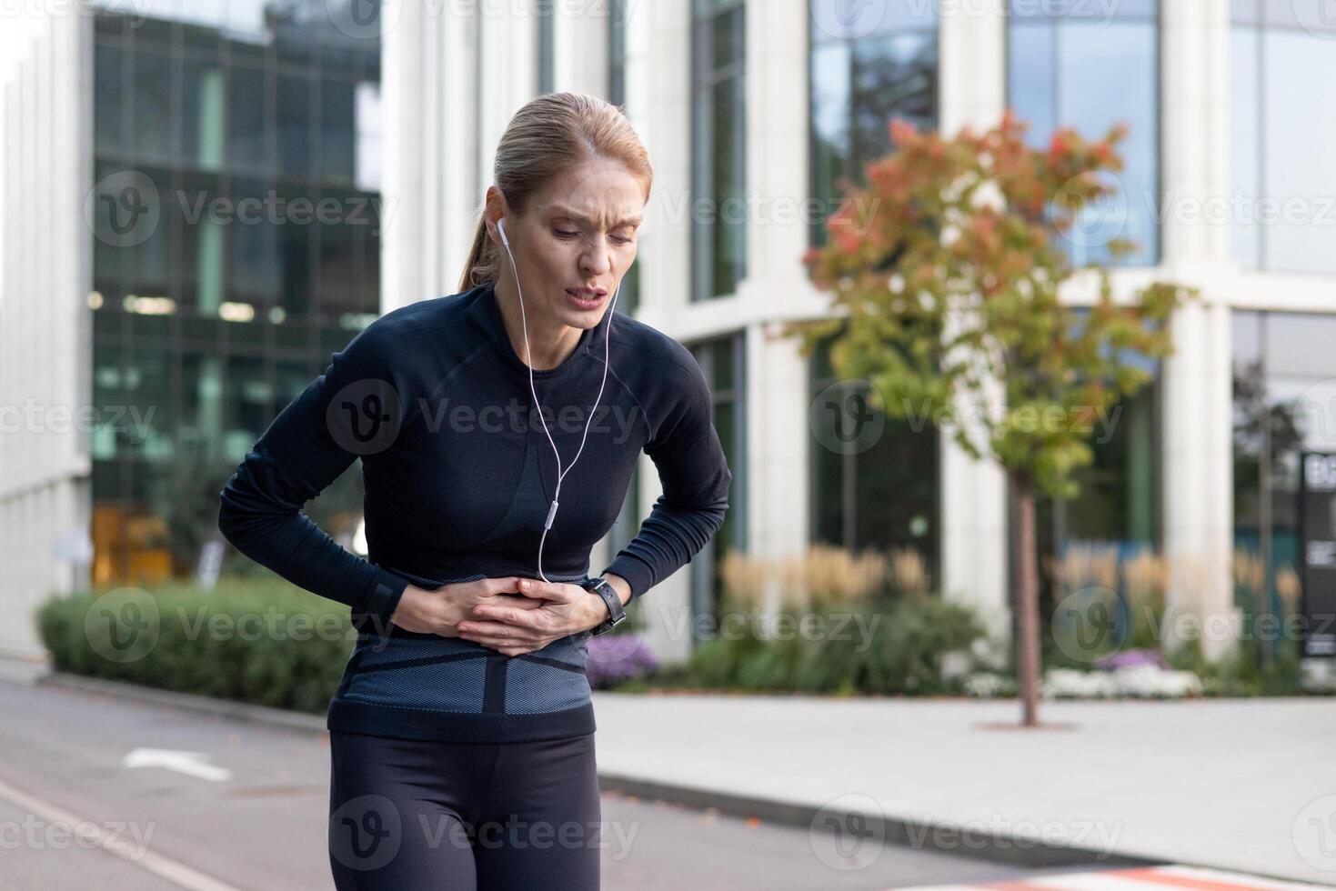Tired overwhelmed woman holding hands on stomach senior mature runner during jogging and fitness class has severe stomach pain bent over massaging muscle. photo