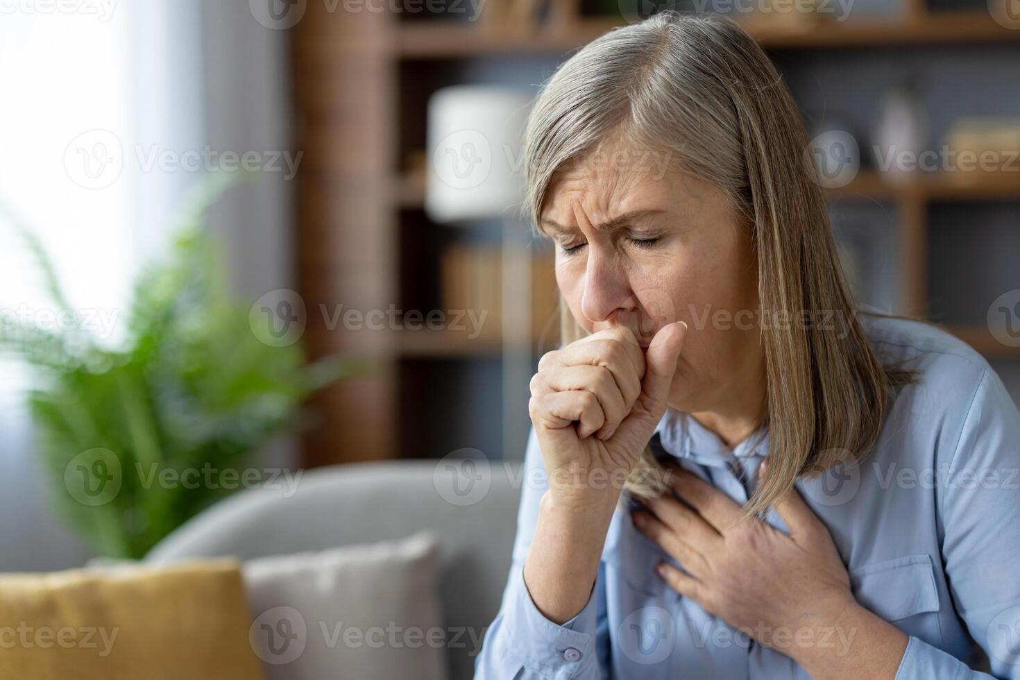 An older woman looks distressed as she coughs, clutching her chest in discomfort. She is sitting in a well-furnished living room, expressing signs of illness. photo