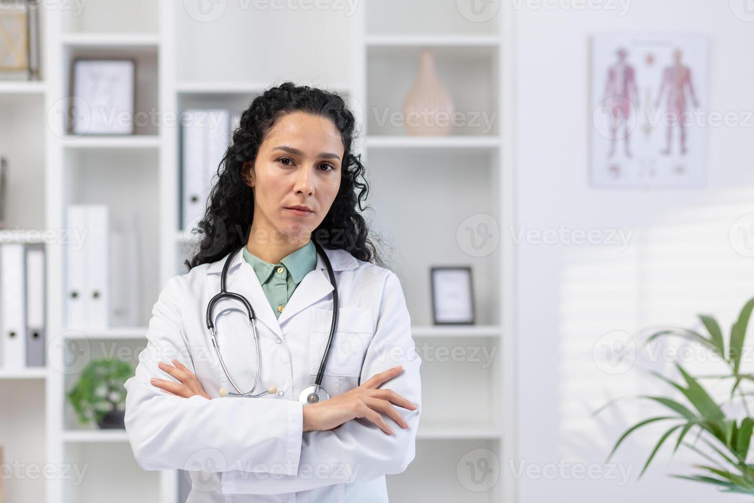 de cerca retrato de grave y confidente Hispano hembra doctor, mujer mirando a cámara con cruzado brazos pensativamente, trabajando a lugar de trabajo dentro clínica oficina, consultante pacientes foto