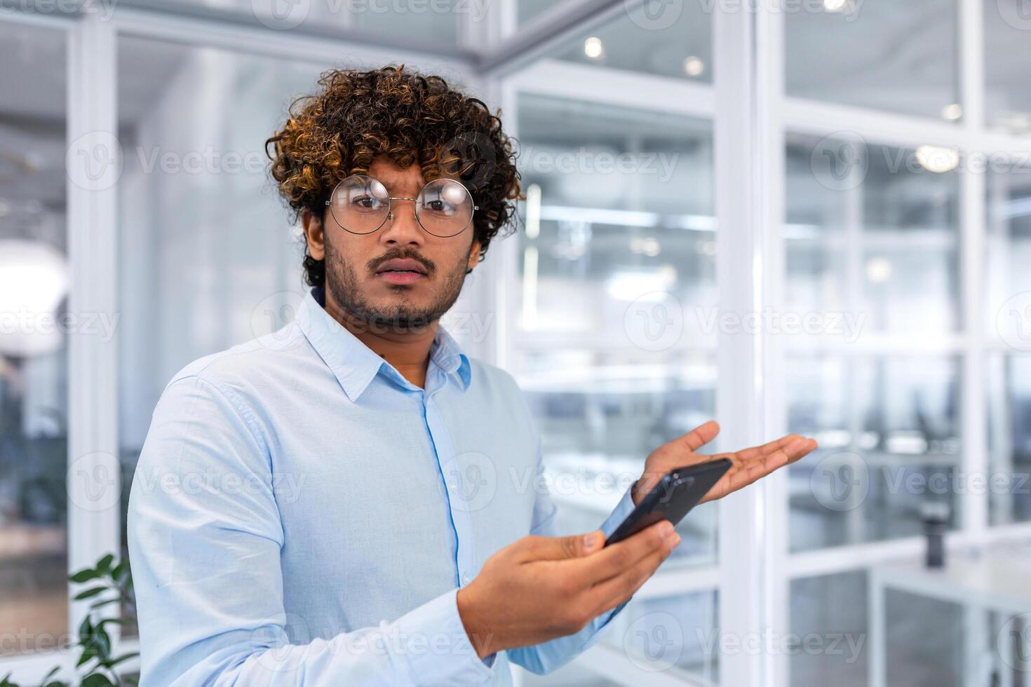 Nervous and unsatisfied man with phone looking at camera, young businessman hindu programmer holding smartphone, got answer bad news and unsatisfactory work results inside office. photo
