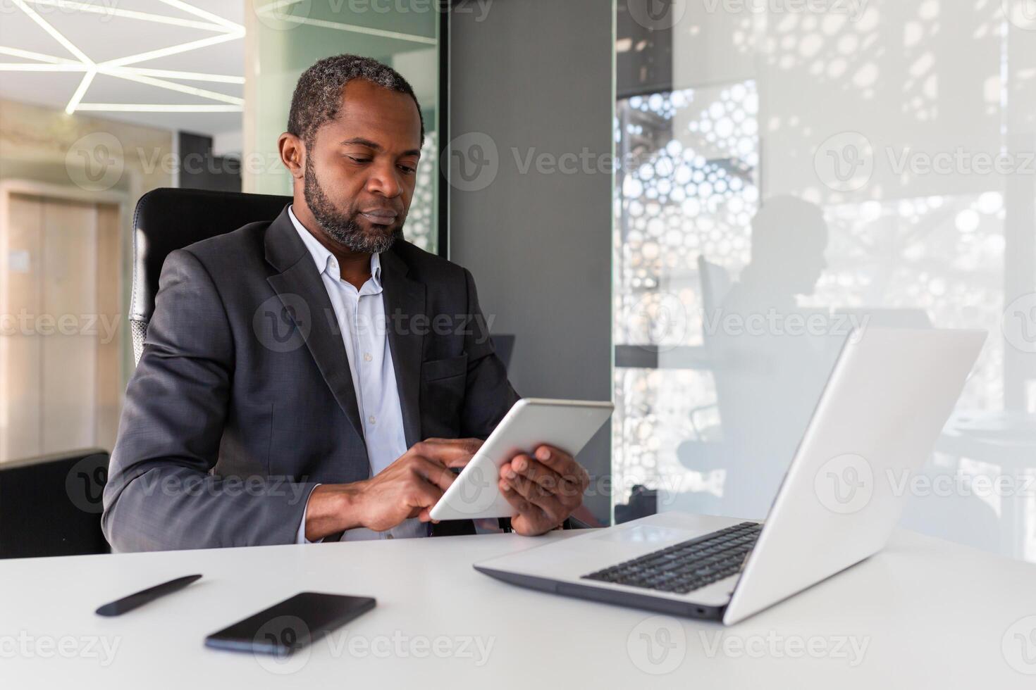 Serious thinking businessman working inside office, using tablet computer, african american mature and confident boss using app at workplace, reading financial performance indicators. photo