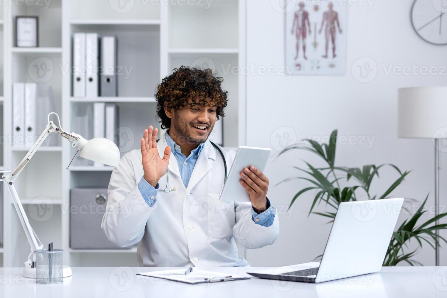 joven exitoso médico utilizando tableta computadora para llamar, hombre en blanco médico Saco clínica trabajador consultante pacientes remotamente sentado a escritorio dentro oficina. foto