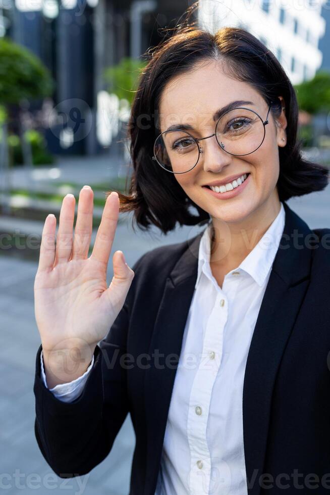 A smiling professional woman in glasses and suit waves at the camera while engaging in a call outside an office building. photo